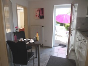 A dining area with a table set for two, a chair, a radiator, a painting on the wall, and an open door leading to an outdoor patio with a pink umbrella.