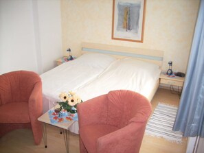 A bedroom with a bed, two chairs, a nightstand, a lamp, and a framed picture on the wall. - VO 2; 1-space; EC; Terrace; 25m² - Gästehaus Vogelsang (Sierksdorf)