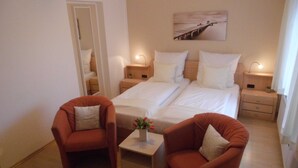 A hotel room with two beds, a small table with a vase, two armchairs, a painting on the wall, and a door leading to a bathroom. - VO 2; 1-space; EC; Terrace; 25m² - Gästehaus Vogelsang (Sierksdorf)