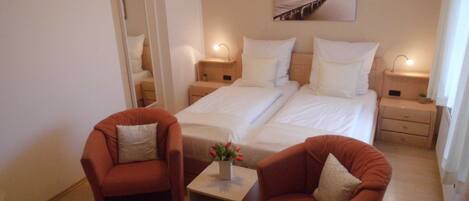 Quarto de hotel com duas camas, uma mesinha com um vaso, duas poltronas, um quadro na parede e uma porta para o banheiro.
