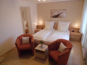 A hotel room with two beds, a small table with a vase, two armchairs, a painting on the wall, and a door leading to a bathroom. - VO 2; 1-space; EC; Terrace; 25m² - Gästehaus Vogelsang (Sierksdorf)