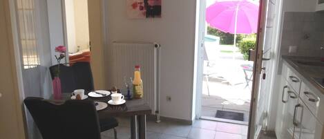 A dining area with a table set for two, a chair, a radiator, a painting on the wall, and an open door leading to an outdoor patio with a pink umbrella.