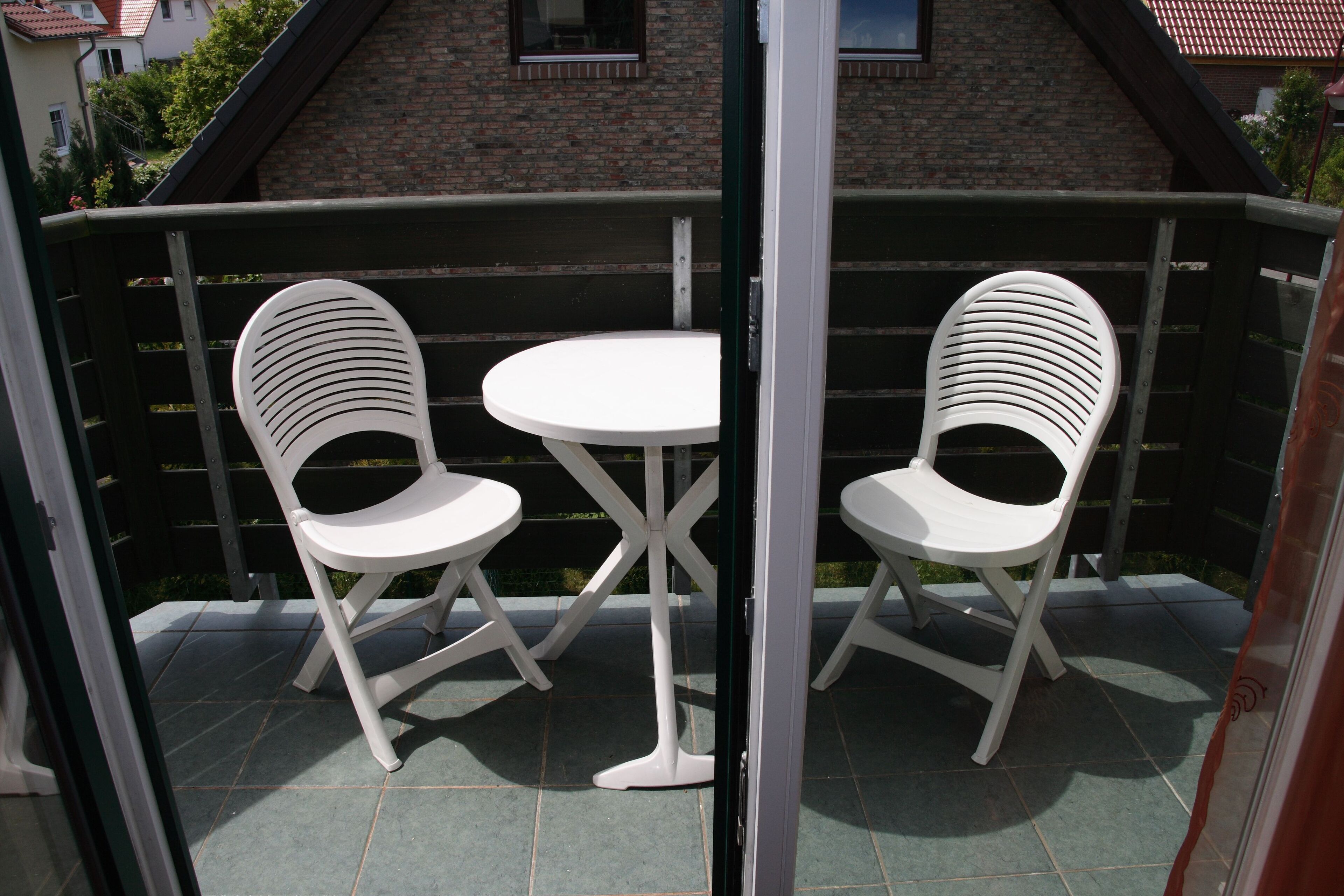 A balcony with two white chairs and a small table, a glass door, and a brick building in the background.