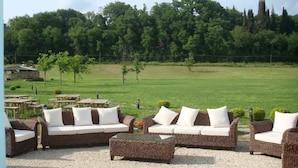 An outdoor seating area with wicker furniture and white cushions, set against a backdrop of a grassy field and trees.
