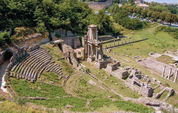 An ancient amphitheater with stone seating and columns, surrounded by greenery and ruins.