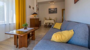 Una sala de estar con un sofá, una mesa de centro de madera, una mesa de comedor con sillas y un cortinón amarillo.