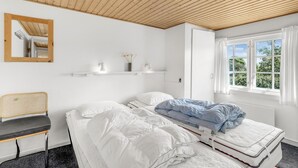 Ein Schlafzimmer mit Holzboden, einem Bett mit weißer und blauer Bettwäsche, einem Sessel, einem Spiegel und einem Fenster mit Blick auf Grünflächen.