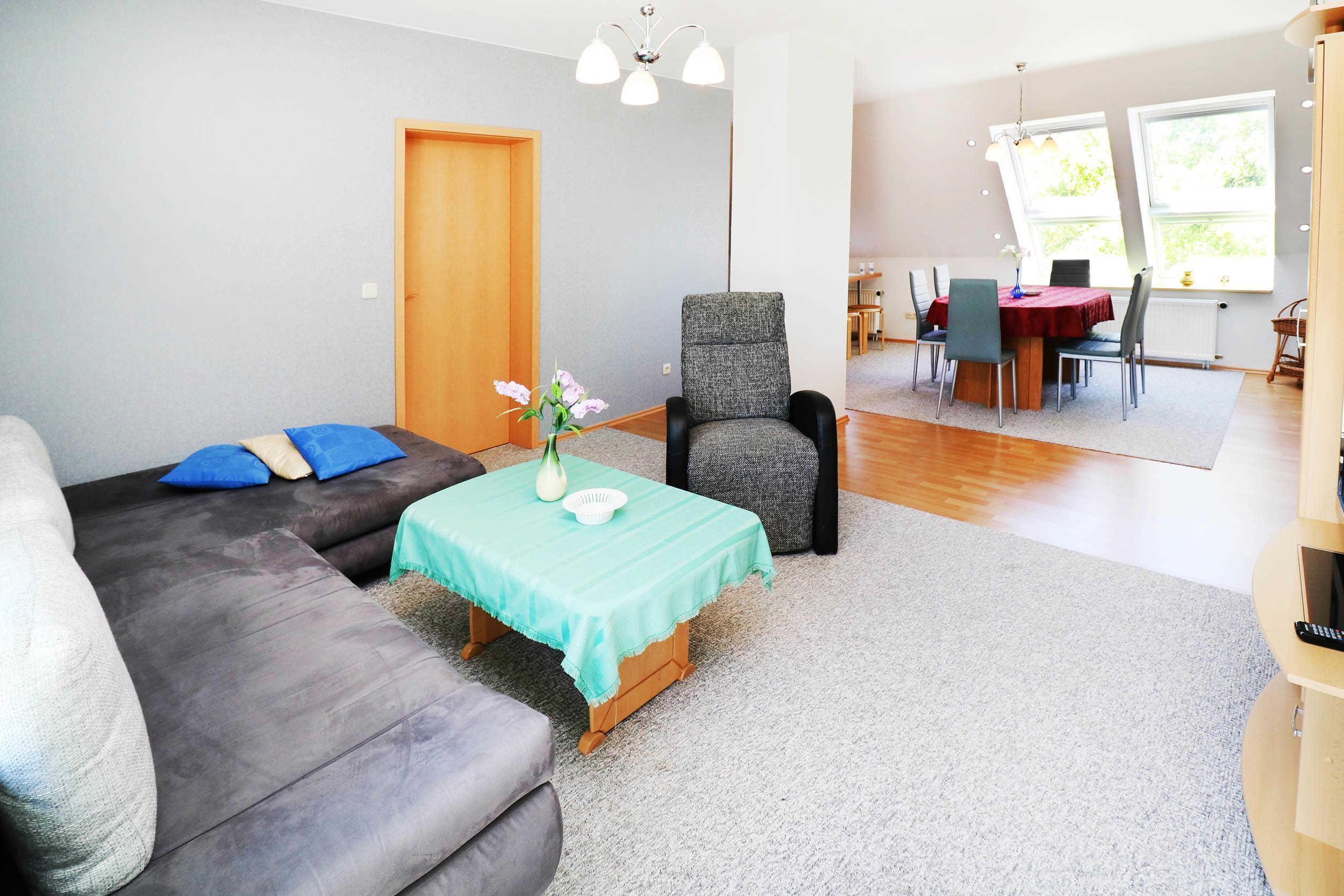 Un salon avec un canapé gris, une petite table recouverte d’une nappe verte et un fauteuil gris. On y trouve une porte en bois et une fenêtre donnant sur un paysage verdoyant.
