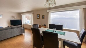 Un salon comprenant un canapé, un téléviseur, une table à manger avec des chaises et de grandes fenêtres donnant sur un plan d’eau.