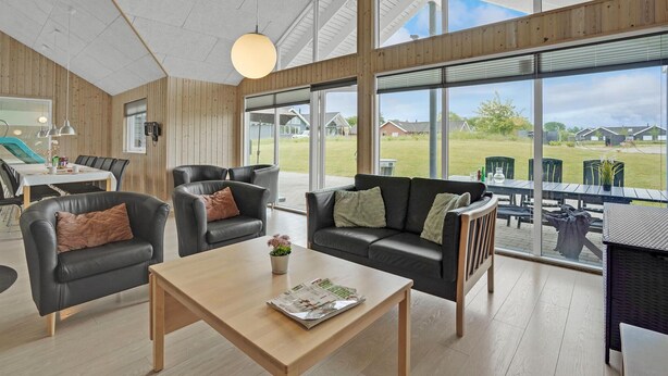 A spacious living area with a wooden floor, a large window offering a view of the outdoors, a dining table with chairs, and a sofa with cushions.