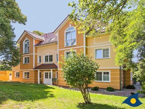 Ein zweistöckiges Haus mit rotem Ziegeldach, einem Hauseingang mit weißer Tür und einem kleinen Vorgarten mit einem Baum.