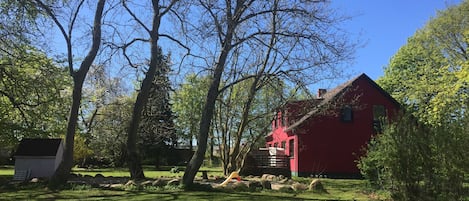 A red house with a sloped roof, surrounded by a grassy yard with trees and a small shed.