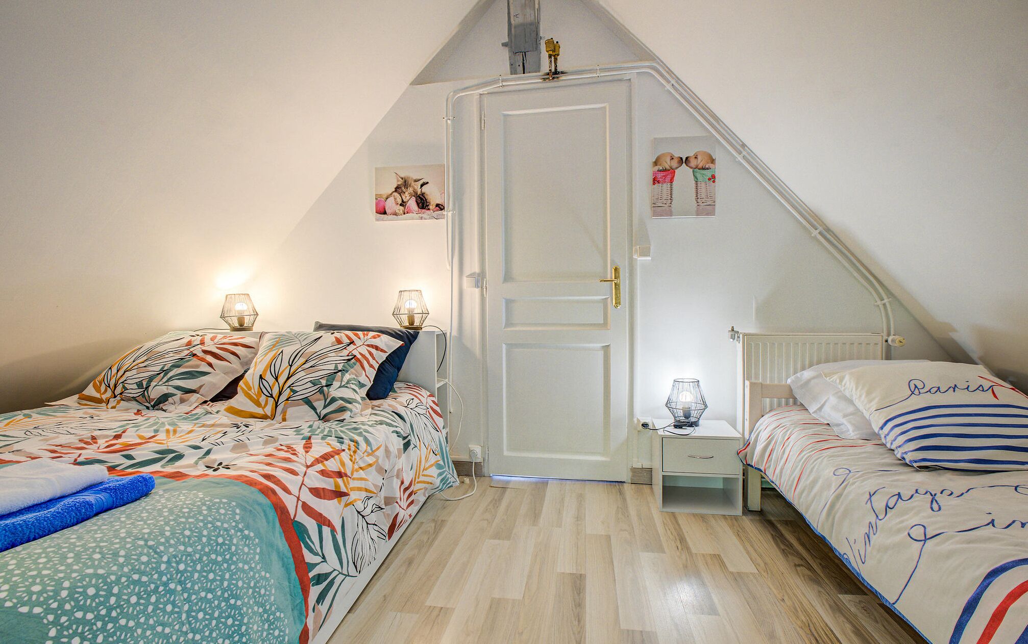 A bedroom with two beds, a white door, a nightstand with a lamp, and a small shelf with decorative items.