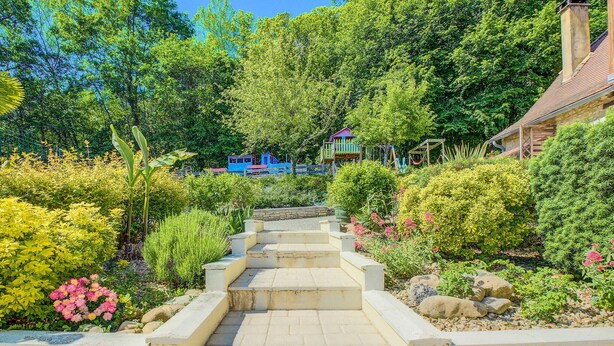 Un sentier de jardin mène à un bâtiment doté d’un toit rouge, qui est entouré d’une végétation luxuriante et de plantes en fleurs.
