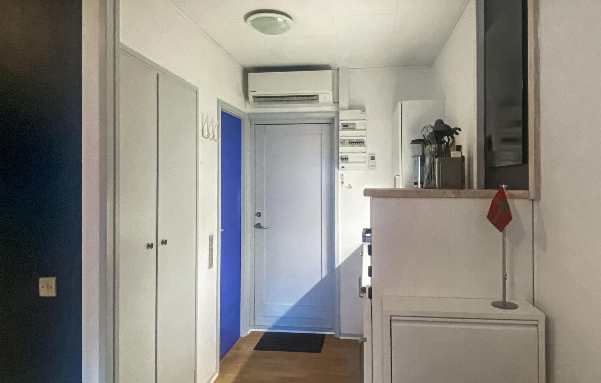 En smal hall med en blå dörr, en luftkonditioneringsenhet som är monterad på väggen och ett litet skåp med ett rött föremål ovanpå.