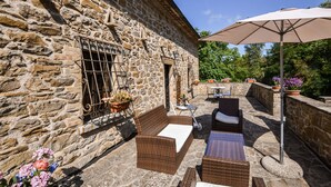 Un edificio de piedra con terraza y mobiliario exterior, incluyendo sillas de mimbre y una mesa, además de un gran paraguas que proporciona sombra.