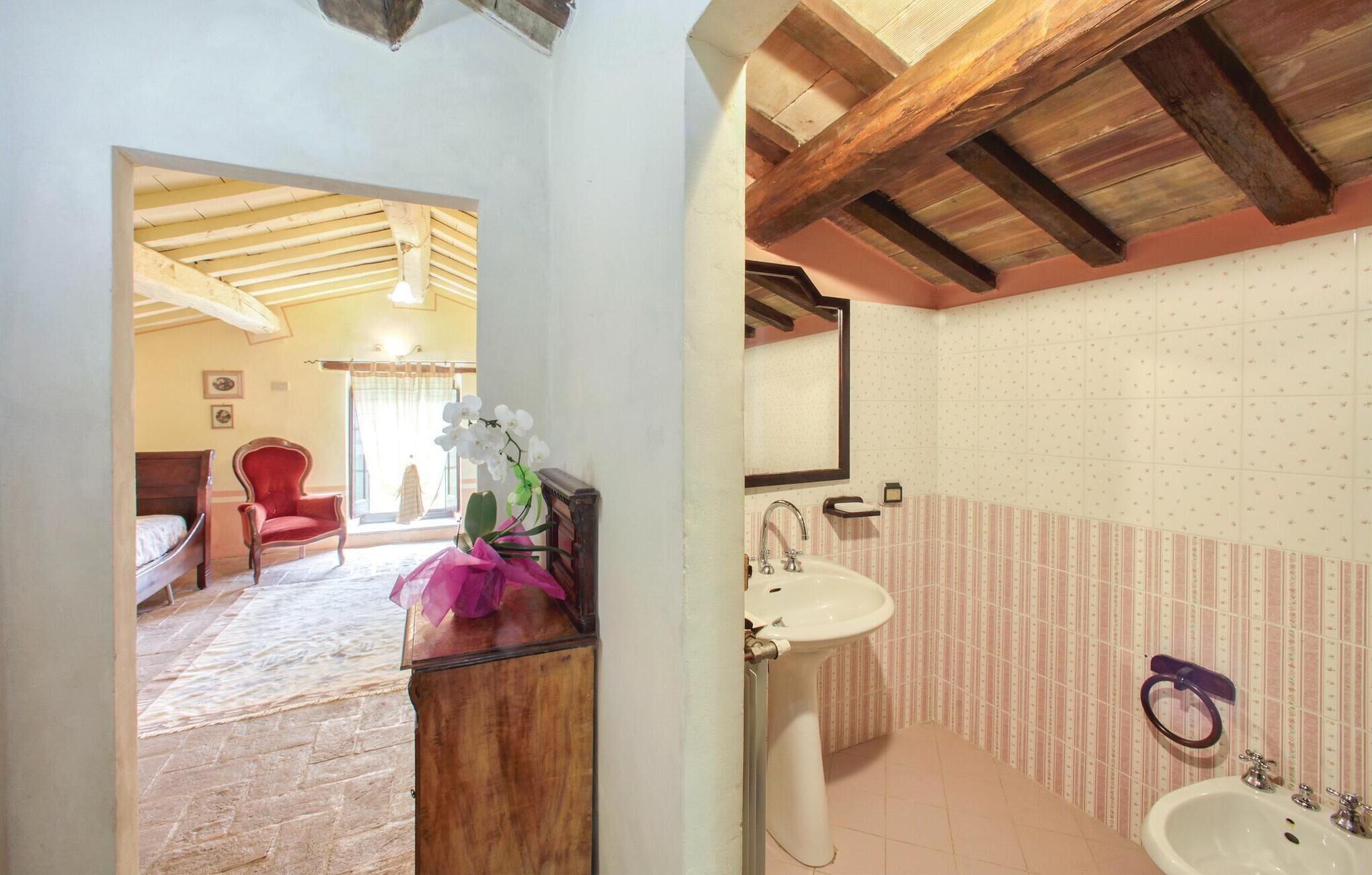 Une salle de bain équipée d’un lavabo, d’un miroir et d’un bidet. La pièce adjacente contient une chaise en bois et un lit.