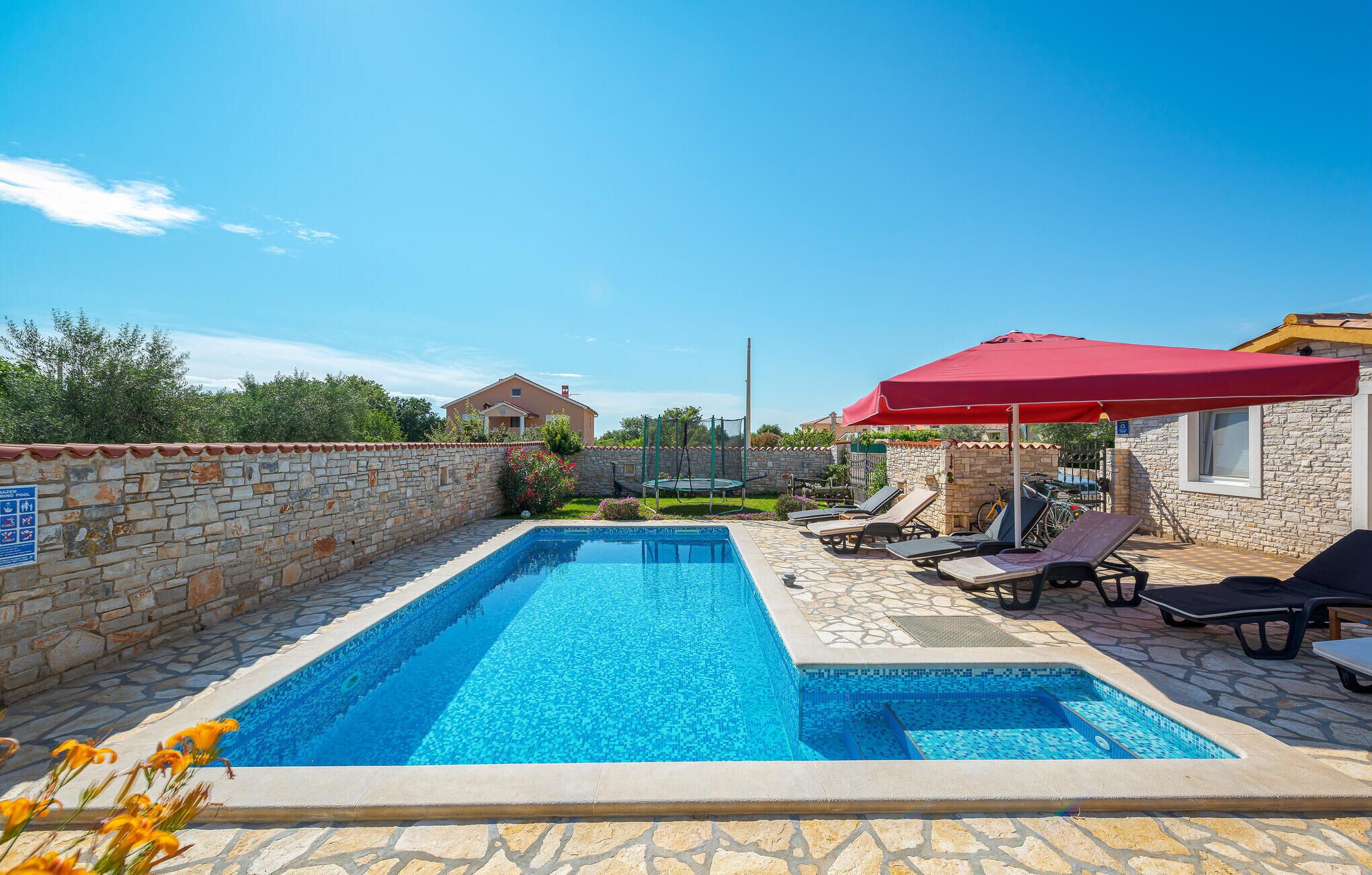 A rectangular swimming pool with a stone border, surrounded by lounge chairs and a red umbrella.