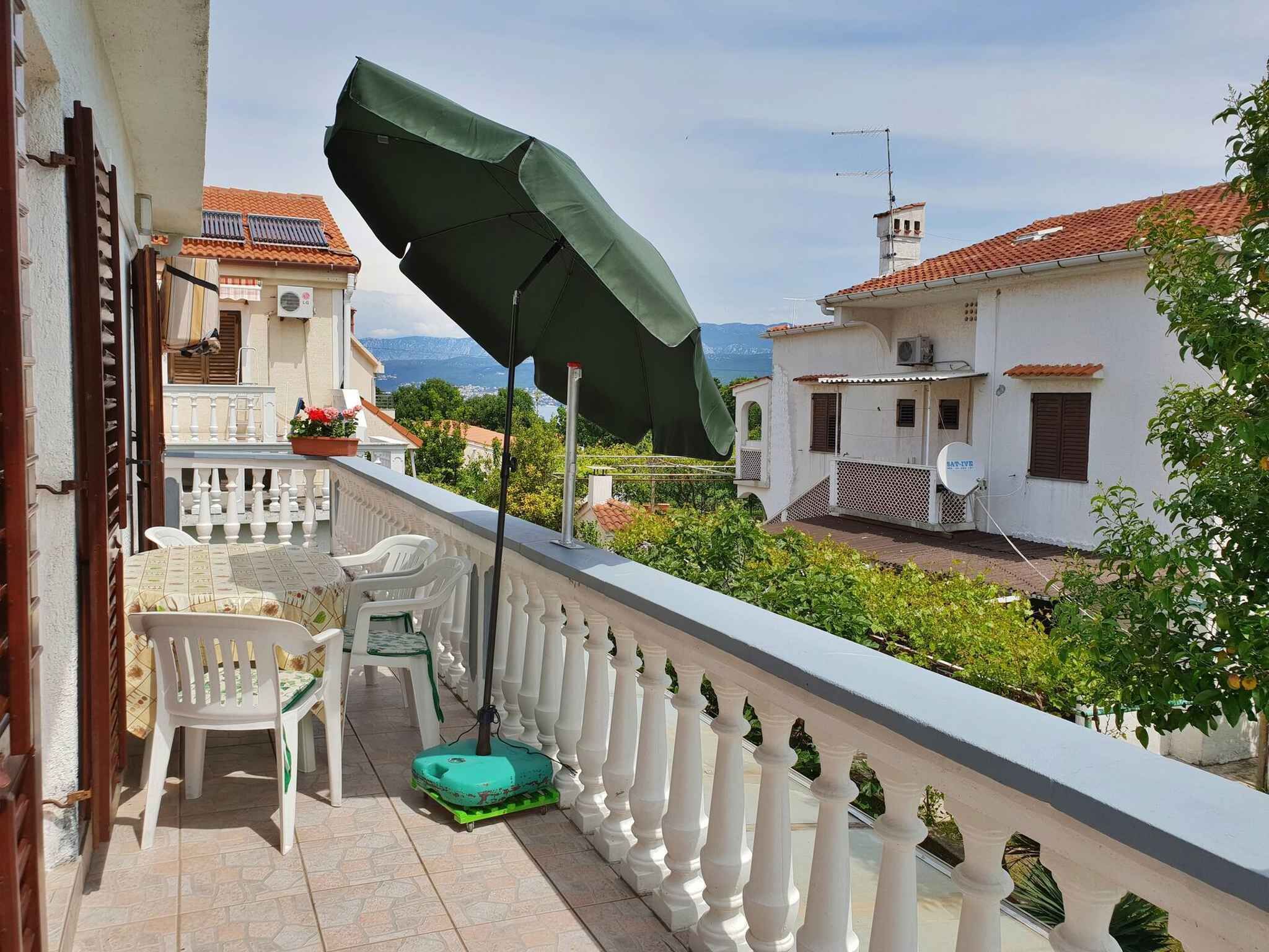 A balcony with white balustrades, a green umbrella, white plastic chairs, and a green exercise mat.