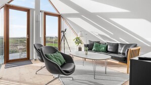 Ein modernes Wohnzimmer mit einem schwarzen Ledersofa, einem schwarzen Sessel mit einem grünen Kissen, einem kleinen runden Tisch und einem großen Fenster mit Blick ins Grüne.