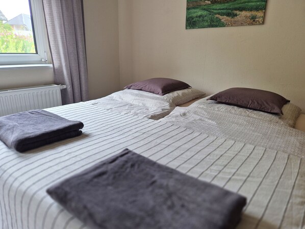 Une chambre d’hôtel avec un lit, des oreillers, une fenêtre avec des rideaux, un radiateur et un tableau au mur.