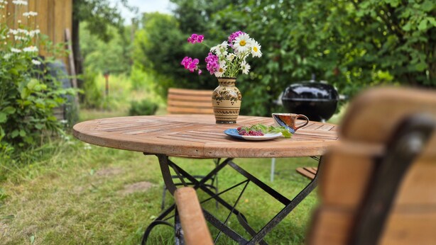 A round wooden table with a metal frame, a vase with flowers, and a grill in a garden setting. - Vacation apartment 2 - Green idyll (Borrentin)