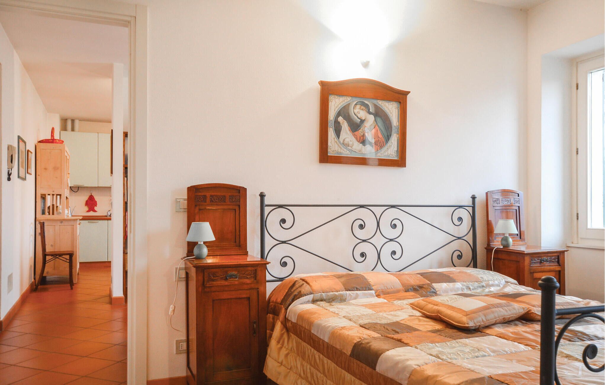 Une chambre à coucher avec un lit, une table de chevet en bois, une lampe, un tableau accroché au mur, et une vue sur une cuisine et une salle de bain par une porte ouverte.