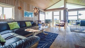 A spacious living area with a leather sofa, a coffee table, and a television. The room has wooden walls and a large window offering a view of the outdoors. - Awesome home in Hejls with sauna (Hejls)