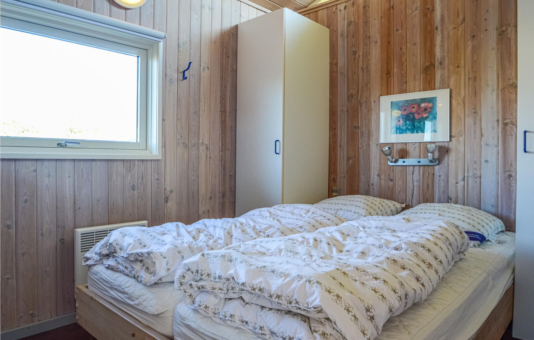 Una camera da letto con parete in legno, una finestra, un letto con biancheria a fiori e un quadro appeso al muro.