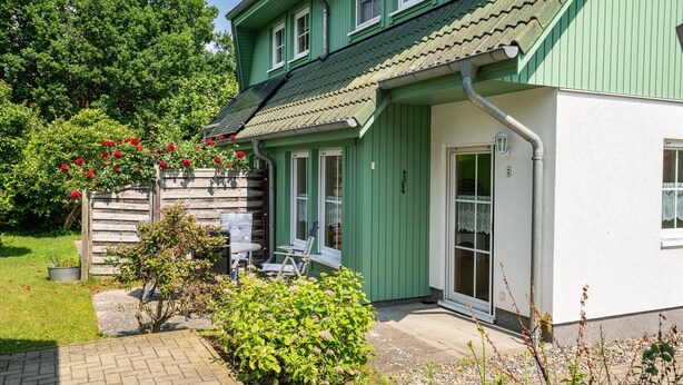 Een groen huis met pannendak, een voordeur en een klein terras met een tafel en stoelen.