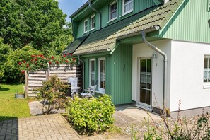 A green house with a tiled roof, a front door, and a small patio area with a table and chairs.