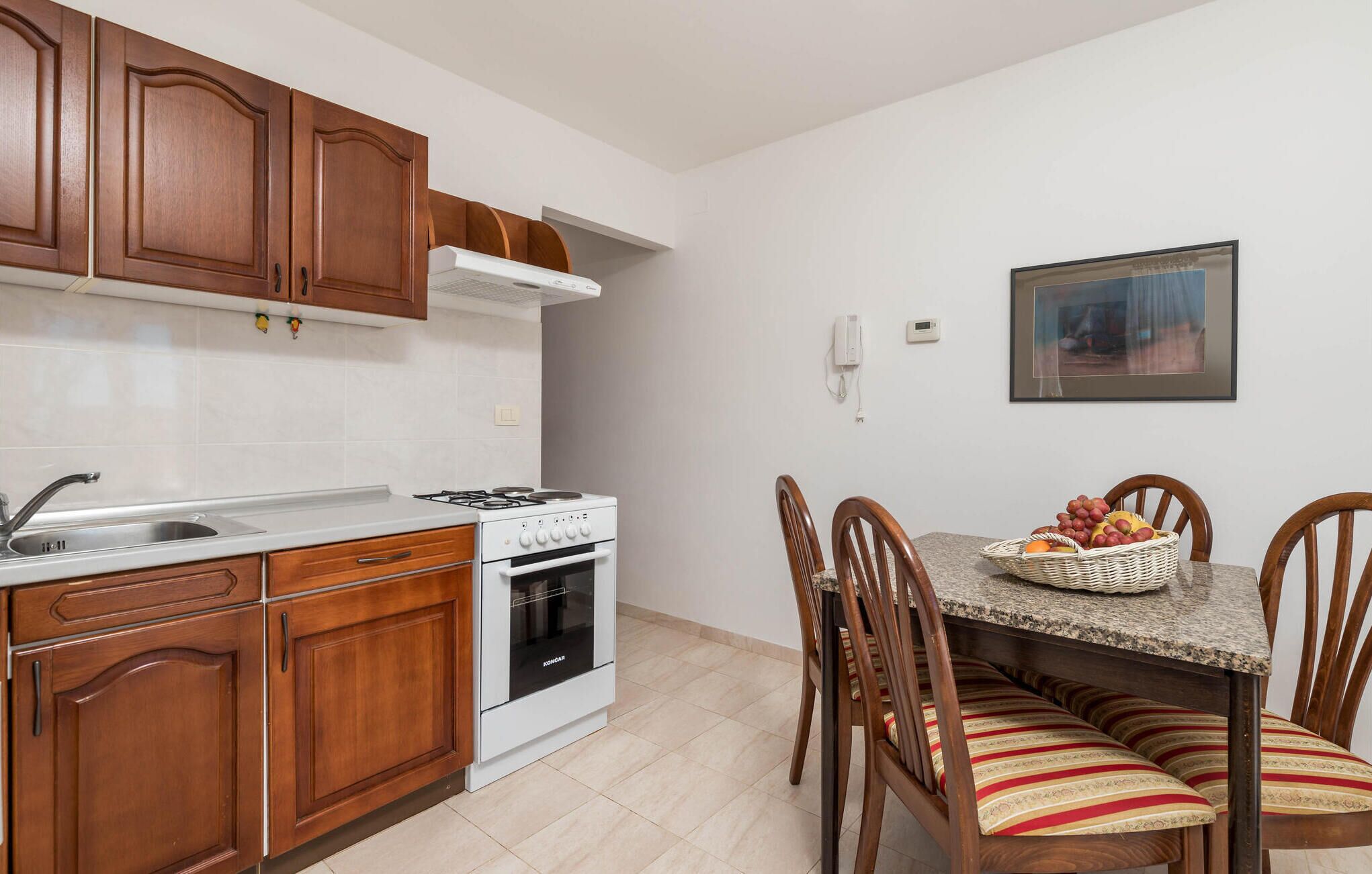 A compact kitchen with wooden cabinets, a gas stove, and a built-in oven. There is a dining table with chairs and a fruit basket.
