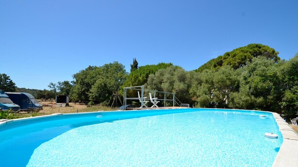 Una piscina azul cristalina con un trampolín y sillas de descanso, rodeada de árboles y vehículos estacionados.