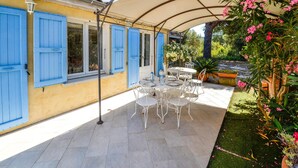 Un patio al aire libre con una mesa y sillas, un área techada con dosel, y un edificio amarillo con contraventanas azules.