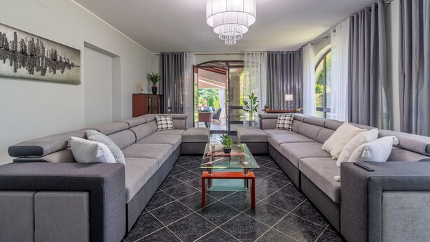 A modern living room with a sectional sofa, a glass coffee table, a chandelier, and a view to an outdoor area.
