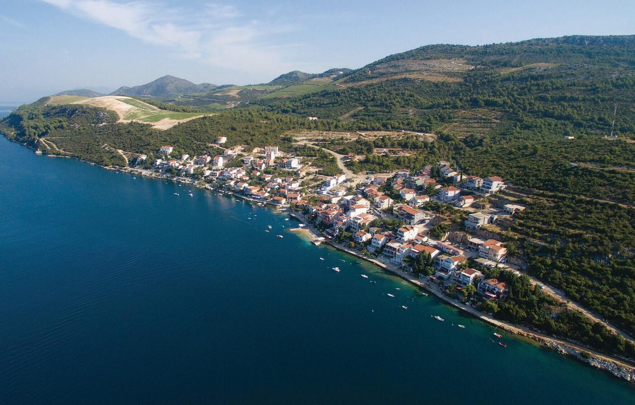 Vista aérea de un pueblo costero con edificios a lo largo de la costa, un mar azul cristalino y terreno montañoso al fondo.
