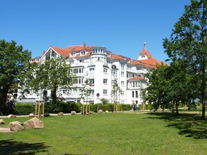 A large white building with red roofs, multiple balconies, and a park in the foreground.
