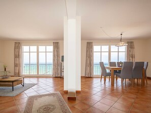 A spacious living area with a dining table, chairs, a TV, and large windows with a view of the sea.