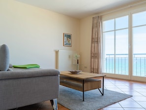 A living room with a sofa, a coffee table, a radiator, a painting, and a view of the sea through a large window. - MB. 4 17 - Ocean View Apt. MB-417 .. (Kühlungsborn)