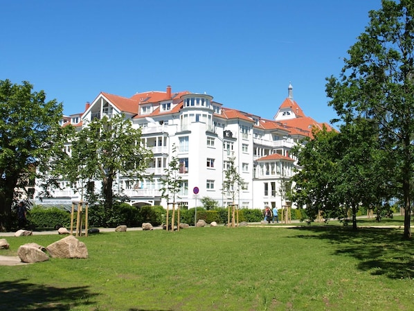 A large white building with red roofs, multiple balconies, and a park in the foreground.