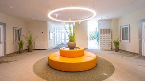 Una hall moderna con un'area salotto circolare centrale, piante in vaso e una lampada a soffitto luminosa.