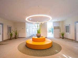 A modern lobby with a central circular seating area, potted plants, and a bright ceiling light.
