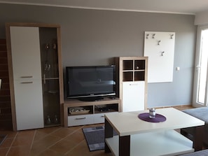Una sala de estar moderna con un televisor de pantalla plana, un mueble de madera, una mesa blanca y un aparador de vidrio con estantes.
