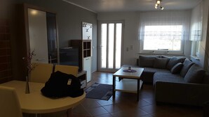 Sala de estar com sofá, TV, mesinha e uma cadeira. Porta deslizante dá acesso a uma varanda.