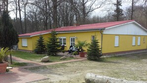 Um edifício amarelo com telhado vermelho, rodeado de árvores e plantas em vasos.