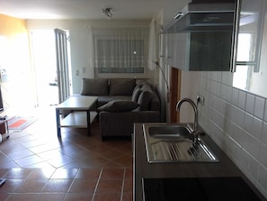 Una cocina compacta con lavamanos integrado, una zona de comedor con mesa y sofá, y un amplio ventanal con cortinas ligeras.