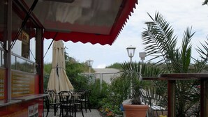 Um café ao ar livre com mesas, cadeiras, toldo vermelho e plantas em vasos.