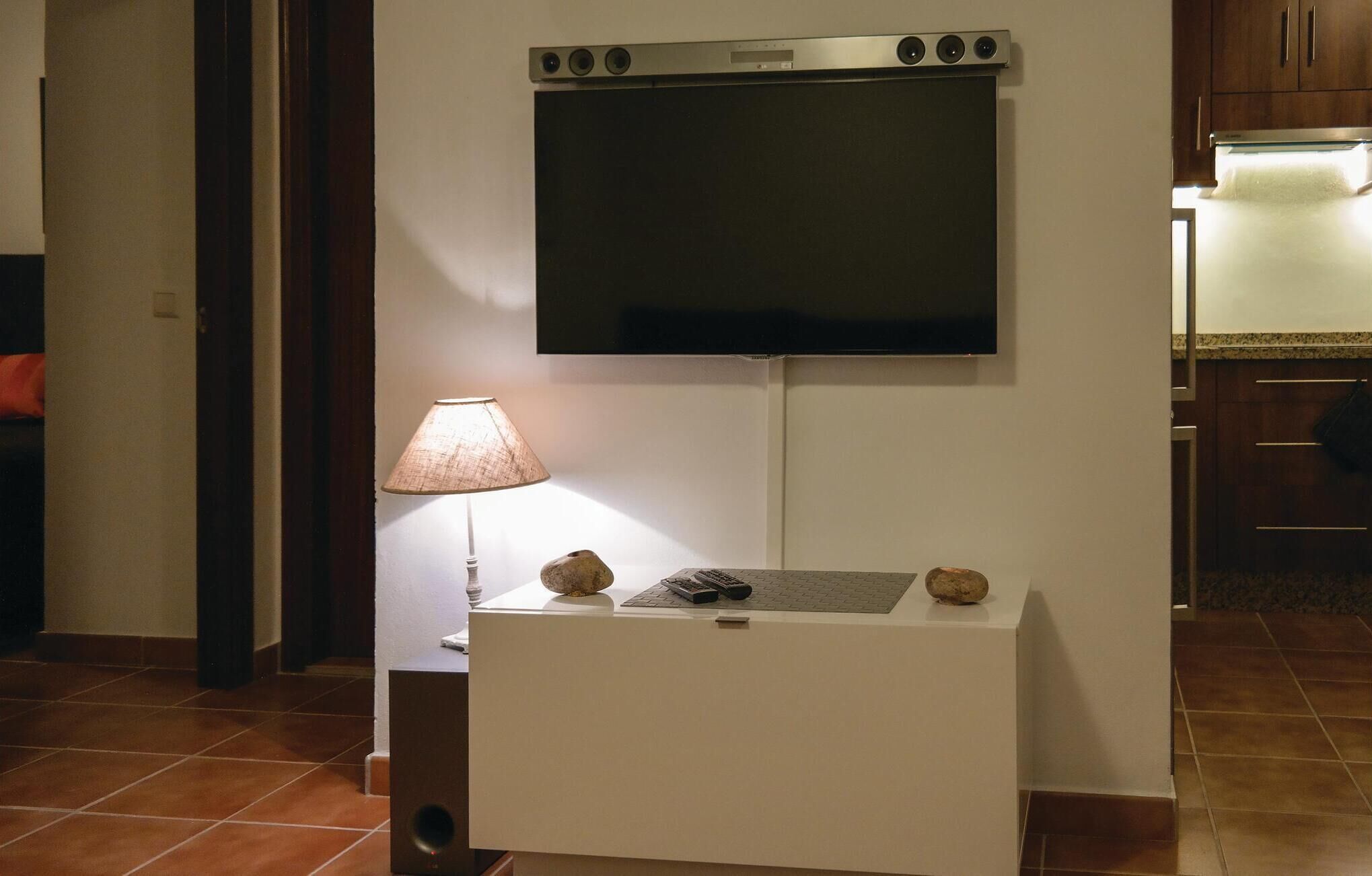 Une pièce dotée d’un téléviseur à écran plat fixé au mur, d’une petite table avec une lampe et des objets décoratifs, et d’un sol carrelé.