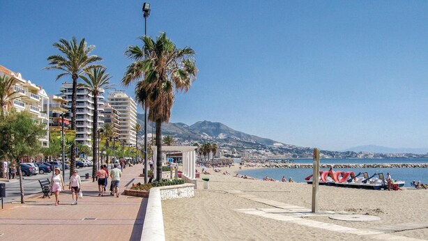 Una passeggiata lungomare con palme, una spiaggia sabbiosa e vista sul mare e sulle montagne in lontananza.