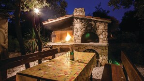 Horno de piedra al aire libre con fuego, rodeado por una mesa y bancos de madera, en un jardín al anochecer.